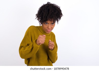 Portrait of attractive Young beautiful African American woman wearing knitted sweater against white wall holding hands in front of him in boxing position going to fight. - Shutterstock ID 1878990478