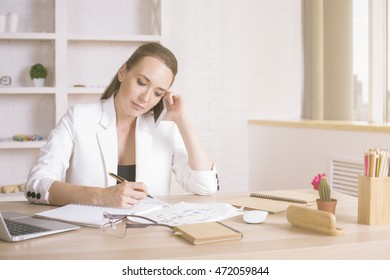 Portrait of attractive young businesswoman talking on phone while doing paperwork at office desk - Shutterstock ID 472059844