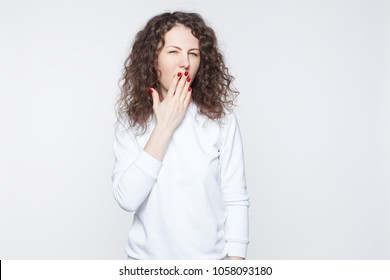 Portrait Of Attractive Young Curly Headed Woman Covering Mouth With Hand, Keeping Secret, Not Supposed To Tell Anything, Looking At Camera With Mysterious Blue Eyes, Posing Isolated Against White Wall