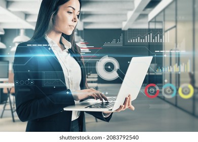 Portrait of attractive young european business woman using laptop with digital business chart interface on blurry office interior background. Finance, trade, technology and future concept.