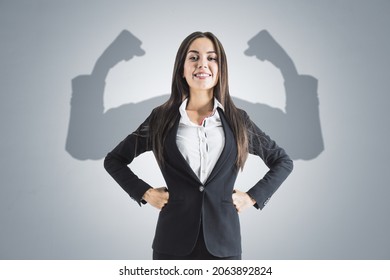 Portrait of attractive young european businesswoman with hands on sides and smile, shadow muscle arms on concrete wall background. Strenght and leadership concept - Powered by Shutterstock