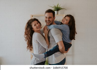 Portrait of attractive young family. The father is standing next to his wife and carrying girl on his back. The family is smiling.