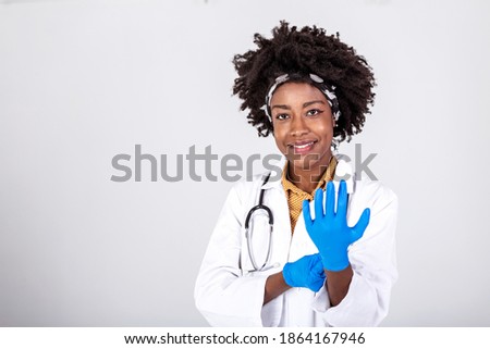Portrait of an attractive young female doctor in white coat putting on medical gloves. Portrait of glad smiling doctor in white uniform standing with crossed hands on gray background