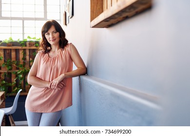 Portrait Of An Attractive Young Female Entrepreneur Smiling Confidently While Leaning Against A Wall By Her Desk In A Large Modern Office