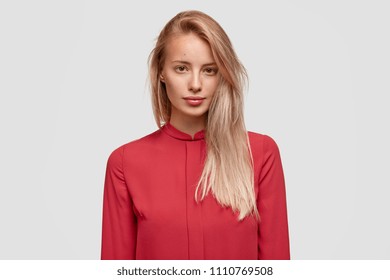 Portrait Of Attractive Young Female With Light Straight Hair, Wears Red Blouse, Poses For Fashion Magazine Against White Studio Wall. Pretty Gorgeous Woman Has Serious Expression, Clean Skin