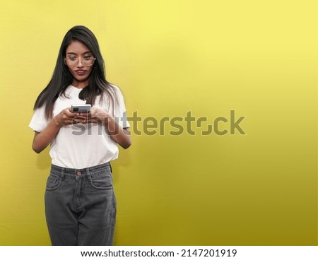 Portrait of an attractive young indian south asian woman or girl using mobile phone while standing with copy space over yellow background
