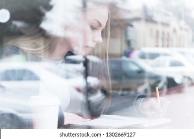 Portrait of an attractive young journalist girl with pen and notepad in cafe behind the showcase. Non-contrast view through window glass reflection