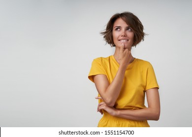 Portrait of attractive young lady with short brown hair looking aside musingly and holding her chin with hand, wearing yellow casual clothes while standing over white background - Shutterstock ID 1551154700