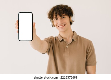 Portrait Of Attractive Young Man In Headphones Holding Smartphone With Blank White Screen And Copy Space, Playlist With Blank Free Space For Mockup