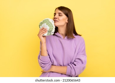 Portrait of attractive young rich woman holding big fan of money, smelling euro banknotes with pleasure, wearing purple hoodie. Indoor studio shot isolated on yellow background.
