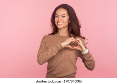 Portrait Of Attractive Young Woman With Brunette Hair Making Heart Shape With Hands And Smiling Friendly To Camera, Showing Symbol Of Love And Care. Indoor Studio Shot Isolated On Pink Background