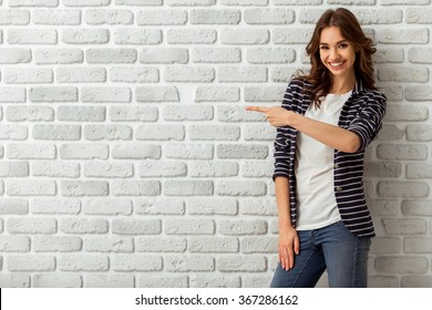 Portrait of an attractive young woman with dark wavy hair, looking into the camera, with white teeth, fingers gestures upwards, on a background of a brick wall