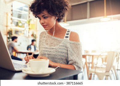 Portrait Of An Attractive Young Woman With Earphones Using Laptop At A Cafe. African American Woman Working On Laptop Computer At A Coffee Shop.