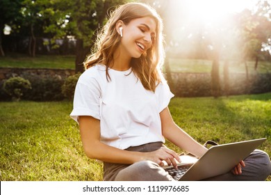Portrait of attractive young woman sitting on green grass in park with legs crossed during summer day while using laptop and wireless earphone