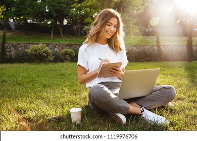 Portrait Of Attractive Young Woman Sitting On Green Grass In Park With Legs Crossed During Summer Day And Writing Notes With Pen While Using Laptop