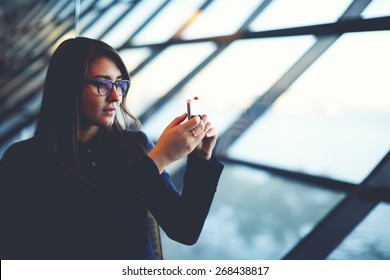 Portrait of attractive young woman taking photo with mobile cell phone sitting near big window of airport hall
