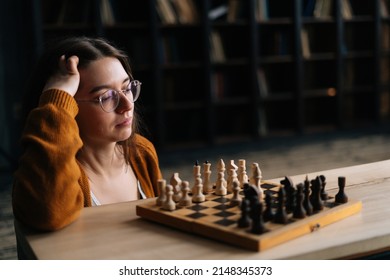 Portrait Of Attractive Young Woman Wearing Elegant Eyeglasses Thinking About Chess Move While Sitting On Wooden Floor In Dark Room. Pretty Intelligent Lady Playing Logical Board Game Alone At Home.