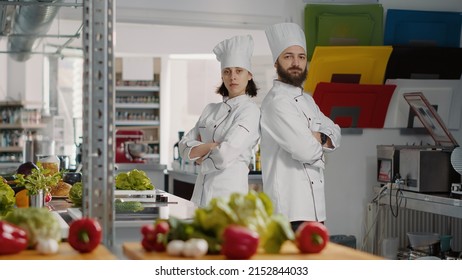 Portrait Of Authentic Cooks Sitting Back To Back In Gourmet Kitchen, Preparing Dish Ingredients. Man And Woman Doing Teamwork Cooking Professional Meal With Gastronomy Cuisine Recipe.