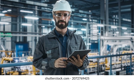 Portrait Of Automotive Industry Engineer In Safety Glasses And Uniform Using Laptop At Car Factory Facility. Professional Assembly Plant Specialist Working On Manufacturing Modern Electric Vehicles.