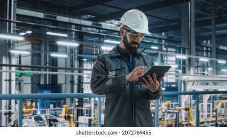 Portrait of Automotive Industry Engineer in Safety Glasses and Uniform Using Laptop at Car Factory Facility. Professional Assembly Plant Specialist Working on Manufacturing Modern Electric Vehicles.