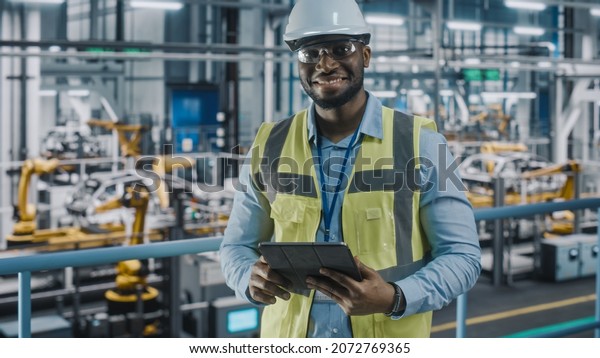 Portrait of Automotive Industry Engineer in\
Safety Uniform Using Tablet at Car Factory Facility. Happy Assembly\
Plant African American Man Specialist Working on Manufacturing\
Modern Electric\
Vehicles.