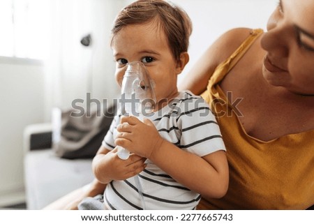 Portrait of a baby boy inhaling medicine in nebulizer with his mother at home.