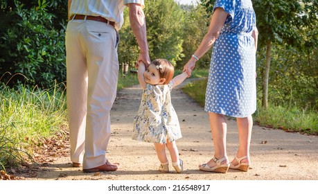 Portrait Of Baby Granddaughter Walking With Her Grandparents On A Nature Path