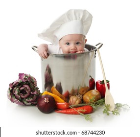 Portrait of a baby sitting wearing a chef hat sitting inside and licking a large cooking stock pot surrounded by vegetables and food, isolated on white