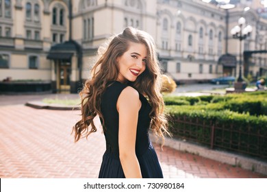 Portrait from back of elegant girl with long curly hair walking on steer on old building background. She has black dress and red lips. She is smiling to camera. - Powered by Shutterstock