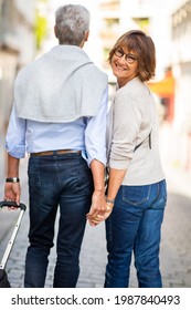 Portrait From Back Of Older Couple Walking On Street With Suitcase. Woman Smiling