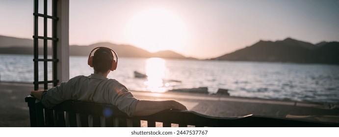 Portrait from the back of a young man in bright big headphones by the sea at sunset. A handsome guy listens to music on the ocean in the rays of the sun, with his back to the camera - Powered by Shutterstock