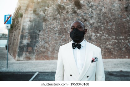 Portrait Of A Bald Bearded Dapper African Guy In A White Suit With A Dotted Bow Tie And A Virus Protective Black Facial Mask, Standing On The Street In Parking Area With A Copy Space Place On The Left