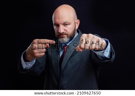 Portrait of a bald business man in suit on dark background. Right hand points to sign boss on his fist. The model is in his 40s, grey and dark hair beard. Showing authority and power concept.