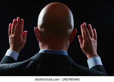 Portrait of a bald businessman in a suit. Back to camera, Hand up in the air. Calm down expression through body language. Dark background.