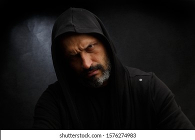 Portrait of a bald man with a beard in a black hood on a dirty gray background.