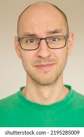 Portrait Of A Bald Young Male In A Green Shirt, With Glasses And With Uneasy Smile - Cringe On A White Background. 