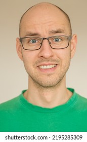 Portrait Of A Bald Young Male In Green Shirt With Glasses And With Uneasy Smile - Cringe On A White Background.