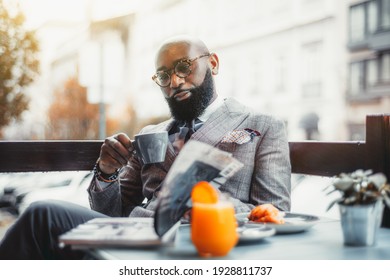 Portrait of a baldheaded adult black dandy guy with a well-groomed beard, in a custom made elegant costume and eyeglasses sitting in a street cafe with a cup of delicious coffee and reading a magazine