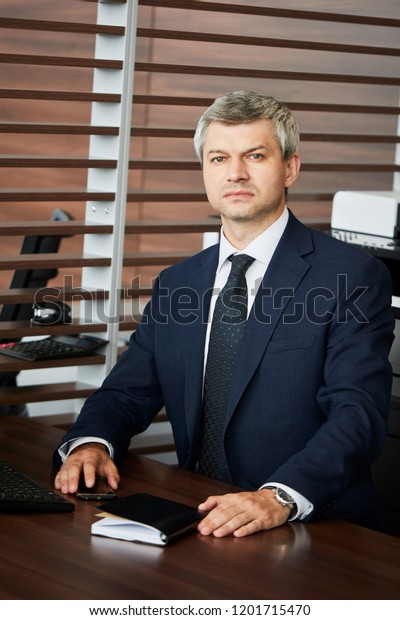 Portrait of\
Banker or successful businessman in blue classy suit, skillful\
sales manager or dealer in suit looking at camera while sitting at\
his office table over auto show\
background