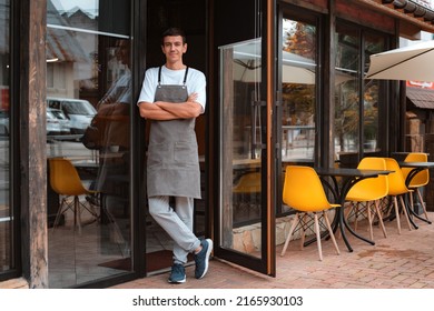 Portrait barista or waiter cafe or coffee shop owner against entrance, gesture inviting you to visit, smiling guy in apron standing outdoors being proud of his small local business - Shutterstock ID 2165930103
