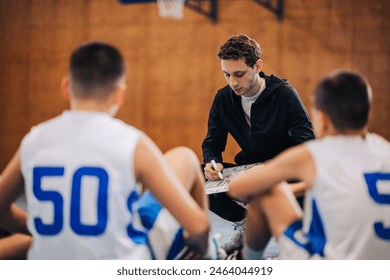 Portrait of a basketball coach crouching at court with his junior team and working on game strategy. A young trainer is drawing on clipboard and working on game strategy and plans with his junior team - Powered by Shutterstock
