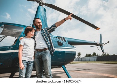 Portrait Of Beaming Bearded Dad Embracing Smiling Child While Gesticulating Hands. They Looking Away While Locating Near Helicopter On Air Stop After Good Flight. Glad Parent Having Fun With Kid