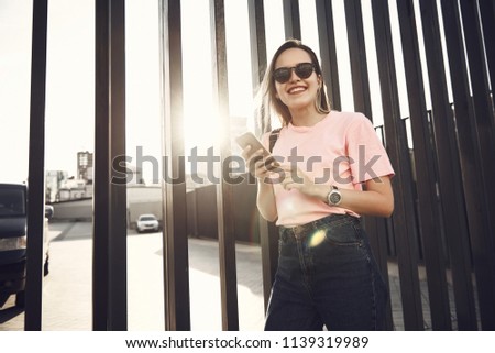 Portrait of beaming girl using modern mobile while posing in urban style. She locating near iron fence on street