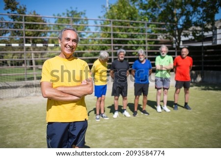 Portrait of beaming senior man on football field. Team leader with gray hair in sport clothes standing, crossing arms, teammates in background. Football, sport, leisure concept