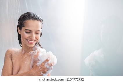 Portrait of beaming woman rubbing body with foam while standing under steam of water. Copy space - Shutterstock ID 1134771752