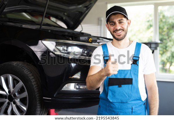 Portrait\
of bearded car mechanic in a car workshop shows thumbs up. Positive\
auto service worker in blue overalls and cap smiling to camera and\
showing thumb up gesture, approving car\
repair