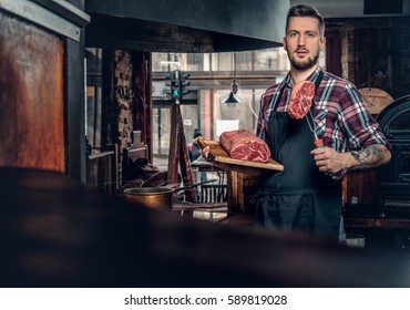 Portrait Of A Bearded Chef With Tattoos On His Arms Hold Beef Steak On A Kitchen.