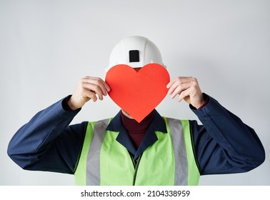 Portrait Of Bearded Construction Site Worker Male Standing By The Wall Holding Red Heart Shape. Worker With Paper Heart In Front Of The Face. Valentine's Day Concept. High Quality Image