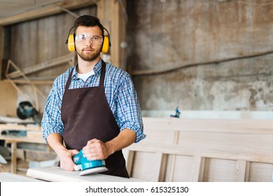 Portrait of bearded craftsman with electric sander in hands distracted from work in order to pose for photography while standing at wooden timber - Powered by Shutterstock