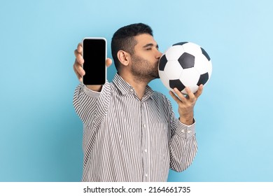Portrait of bearded handsome man standing kissing soccer ball and showing mobile phone with empty black display, wearing striped shirt. Indoor studio shot isolated on blue background.